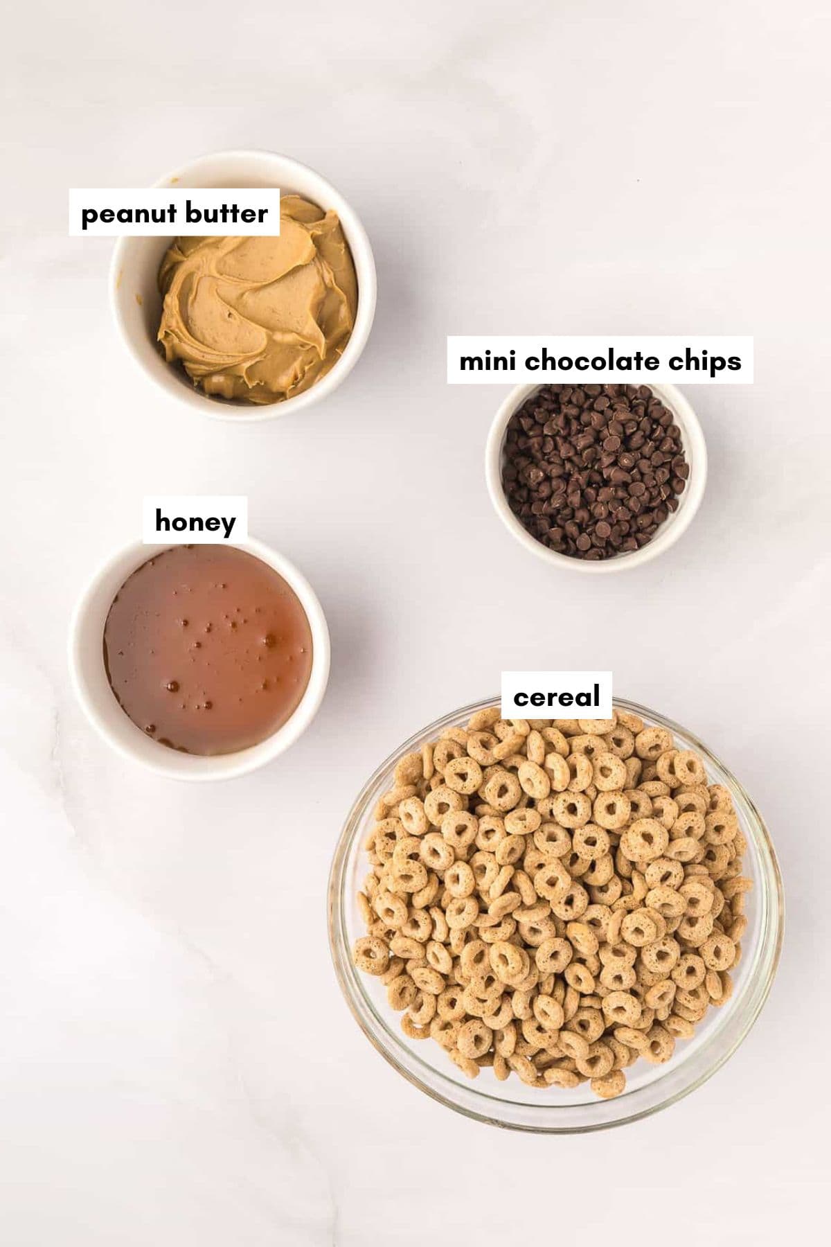 Four bowls on a white surface containing peanut butter, mini chocolate chips, honey, and cereal.