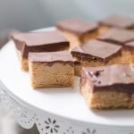 No Bake Buckeye Bars with a thick peanut butter base and rich chocolate topping are arranged in perfect squares on a white, decorative cake stand.
