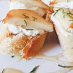 Close-up of sliced baguette topped with a layer of goat cheese, pear slices, fresh herbs, and drizzled with honey, creating a delectable goat cheese crostini on a white plate.