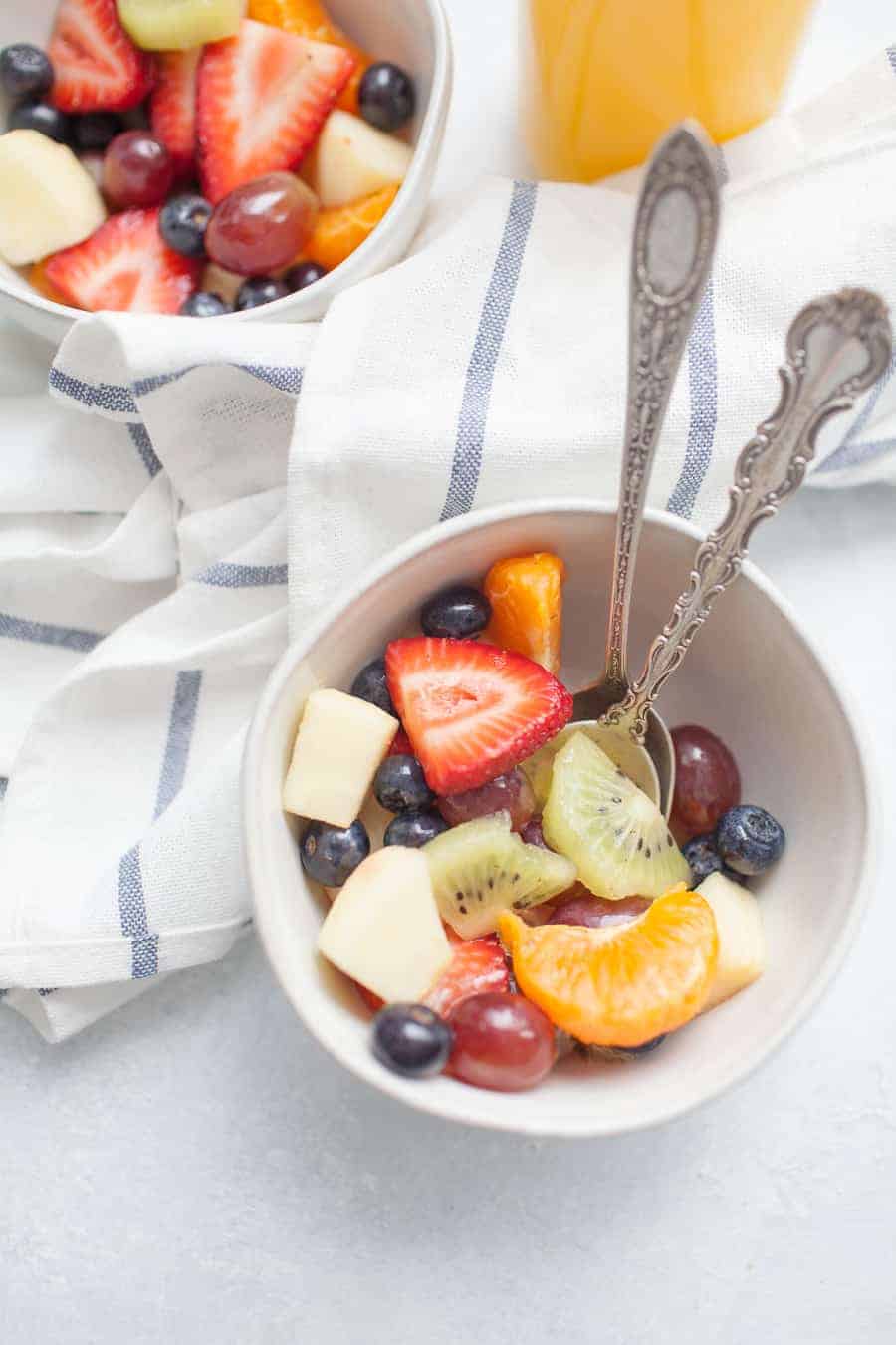 fruit salad in white bowls with silver spoons next to a jug of orange juice 