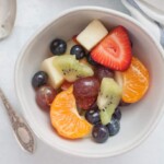 A bowl of Fresh Fruit Salad including strawberries, kiwi, orange slices, grapes, blueberries, and apple pieces, with a spoon beside it.
