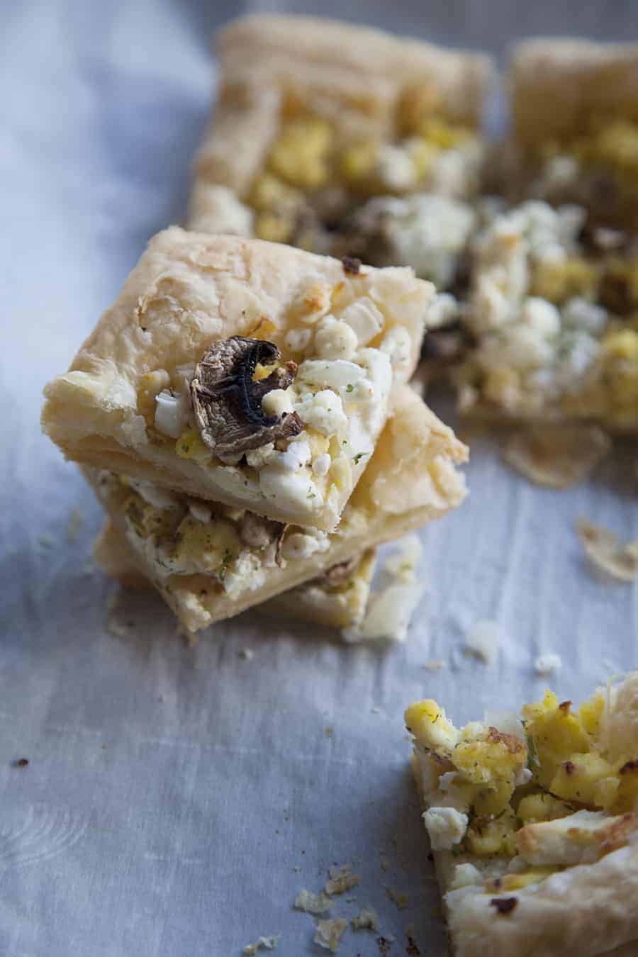 A slice of egg and mushroom tart topped with a flaky golden breakfast puff pastry, delicately placed on a piece of paper.