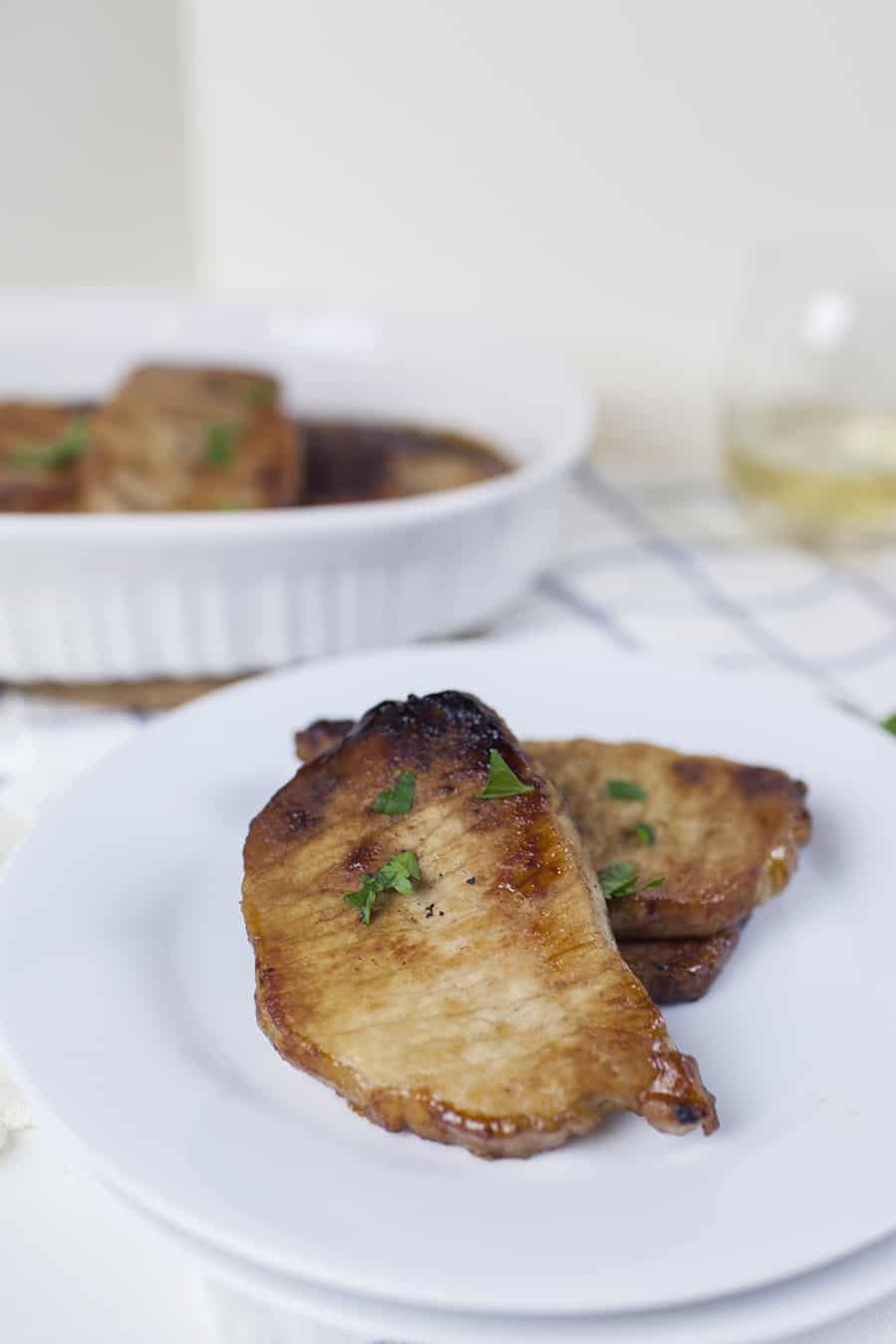 Pork chops on a white plate next to a glass of wine. The white plate beautifully showcases the succulent pork chops, cooked to perfection. Beside it sits a glass of wine, inviting