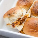 Three chicken sliders with shredded chicken fill soft slider buns in a white dish, topped with herbs and lightly browned on top.