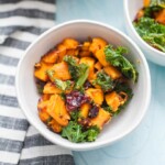 A white bowl filled with sweet potato kale, roasted to perfection and mixed with dried cranberries, sits on a blue surface next to a striped cloth.