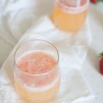 Two glasses of light pink, frothy Bellini recipe-inspired beverage sit on a white cloth, with fresh strawberries partially visible in the background.