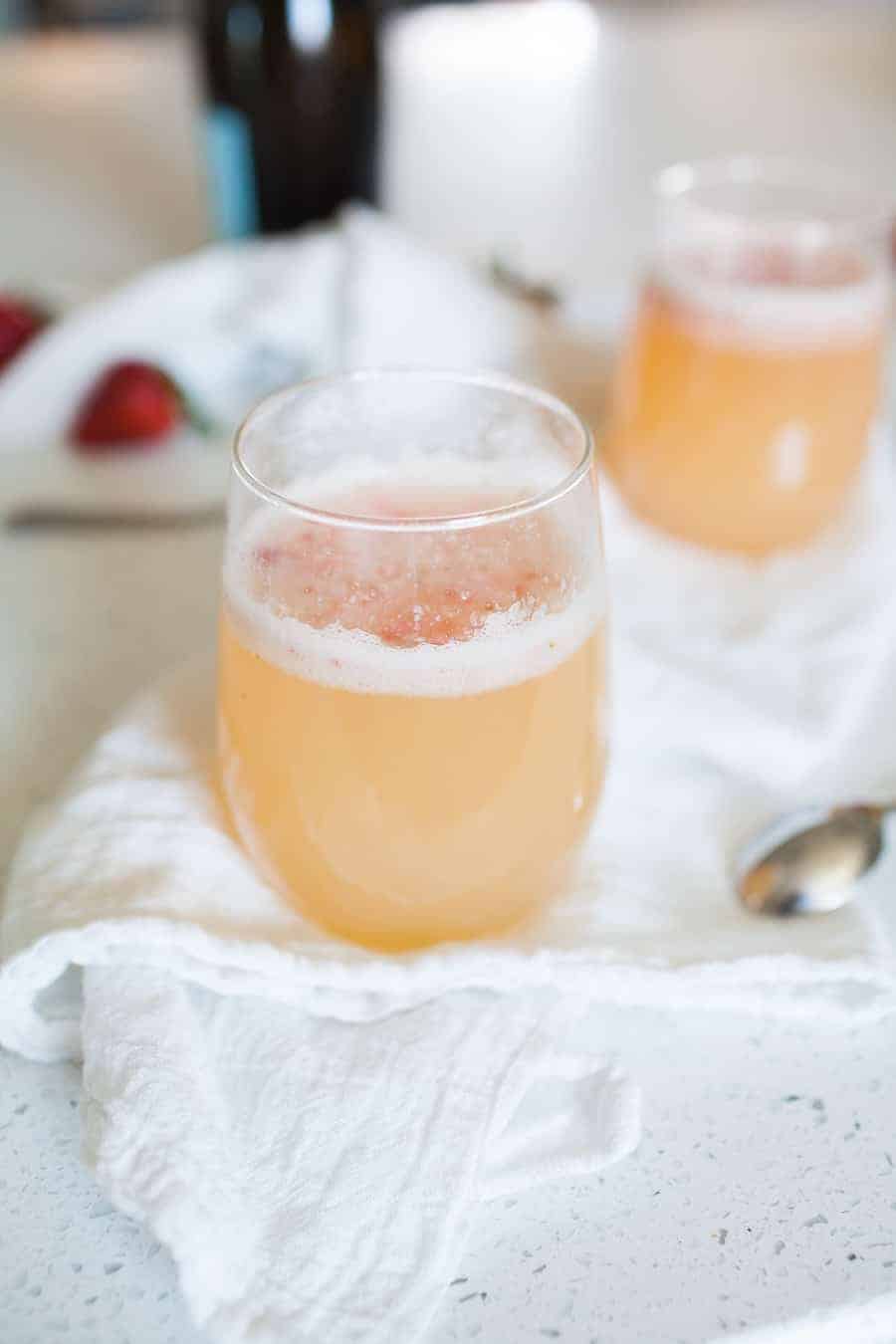 A close-up of a stemless glass filled with a frothy light orange Bellini recipe, placed on a white cloth with a spoon nearby. Another similar drink and strawberries are blurred in the background.