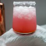 A glass of pink ice-filled prickly pear margarita with a sugar-rimmed edge is placed on a white cloth.