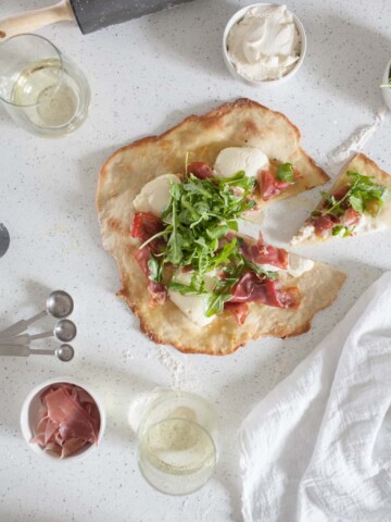 Overhead photo of prosciutto and ricotta thin crust pizza on the counter with small bowls filled with prosciutto and ricotta.