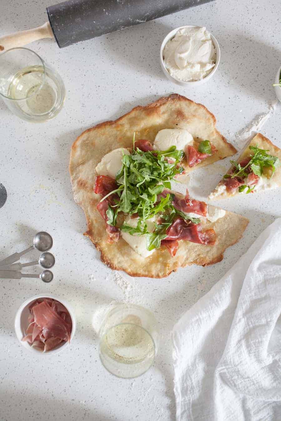 Overhead photo of prosciutto and ricotta thin crust pizza on the counter with small bowls filled with prosciutto and ricotta.