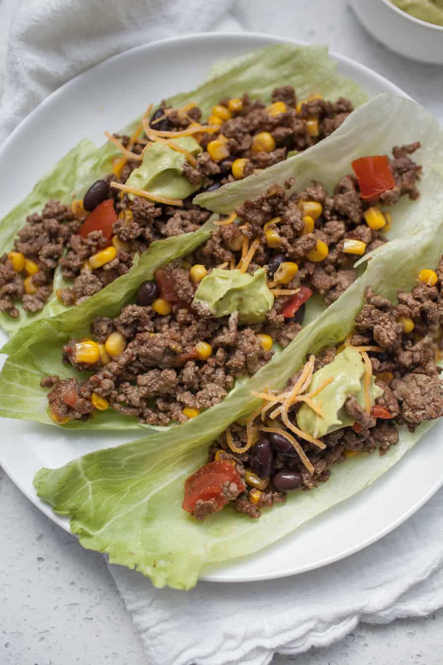 Three beef lettuce wraps filled with ground beef, corn, black beans, tomato, cheese, and avocado are elegantly arranged on a white plate.