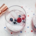 A chilled glass of White Wine Spritzer garnished with blueberries and a raspberry, placed on a white napkin with blue and red stars.