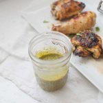A jar of green sauce is in the foreground, with three pieces of cooked seasoned chicken on a white plate in the background, each infused with a delightful cilantro lime marinade.
