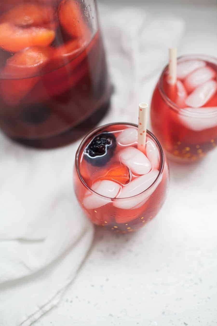 Two glasses of iced blackberry lemonade with straws and berries sit beside a large pitcher on a light surface.