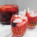 Two glasses of iced red fruit punch with berries and straws sit on a white surface; a large pitcher of blackberry lemonade is in the background.
