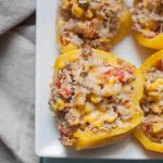 Four yellow bell peppers, reminiscent of classic quinoa stuffed peppers, are filled with a savory mix of ground meat, corn, rice, and melted cheese and arranged on a white rectangular plate next to a beige cloth napkin.