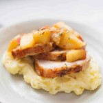 Slices of roasted pork medallions topped with cooked apples and herbs, served on a bed of mashed potatoes on a white plate.