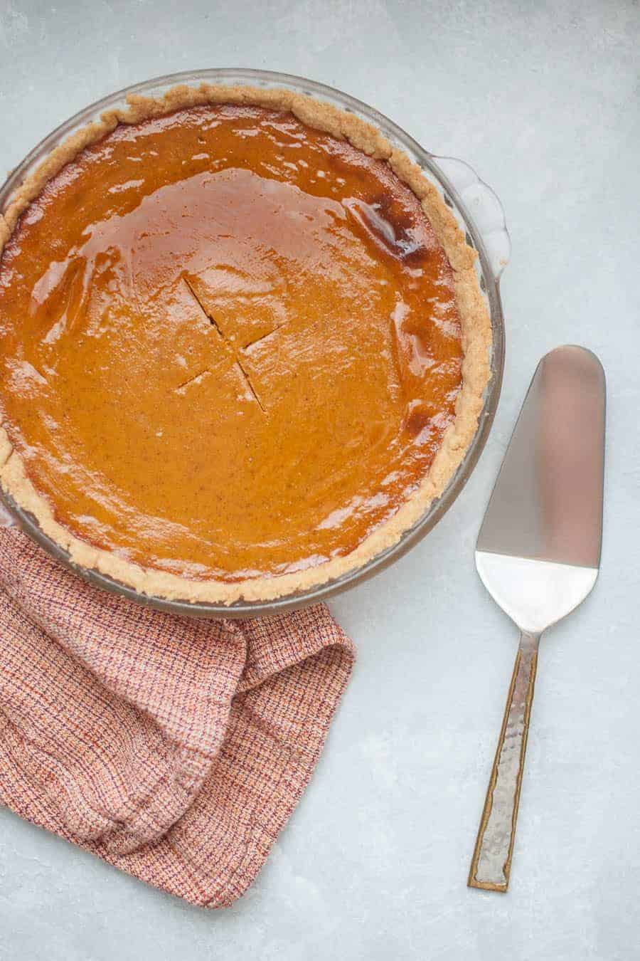 a pumpkin pie in a glass baking dish with a pie server and an orange towel