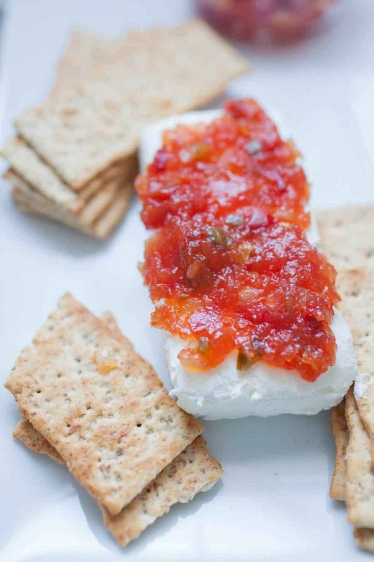 Pepper jelly is poured over top a block of cream cheese next to a plate of crackers. 