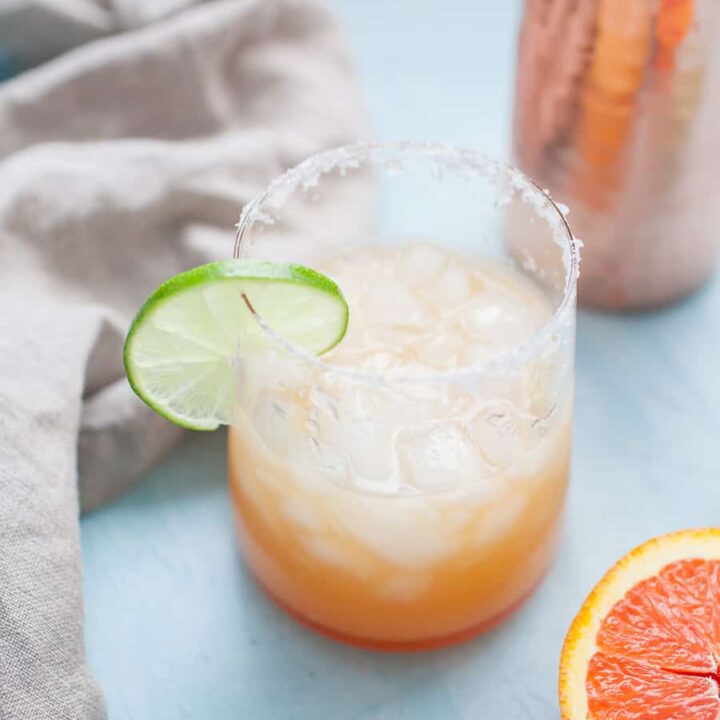 A refreshing orange margarita with a salted rim is garnished with a lime slice, set on a light blue surface beside a half grapefruit, a cloth, and a copper shaker.
