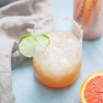 A glass of an iced orange margarita with a salted rim, garnished with a lime wedge, sits next to an orange slice and a beige napkin.