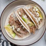 Three pulled pork tacos filled with shredded meat, avocado slices, and crumbled cheese are artfully arranged on a round plate, accompanied by a striped cloth.