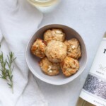 A bowl of baked chicken meatballs beside a bottle of white wine on a white surface, adorned with a sprig of rosemary and a white linen cloth.