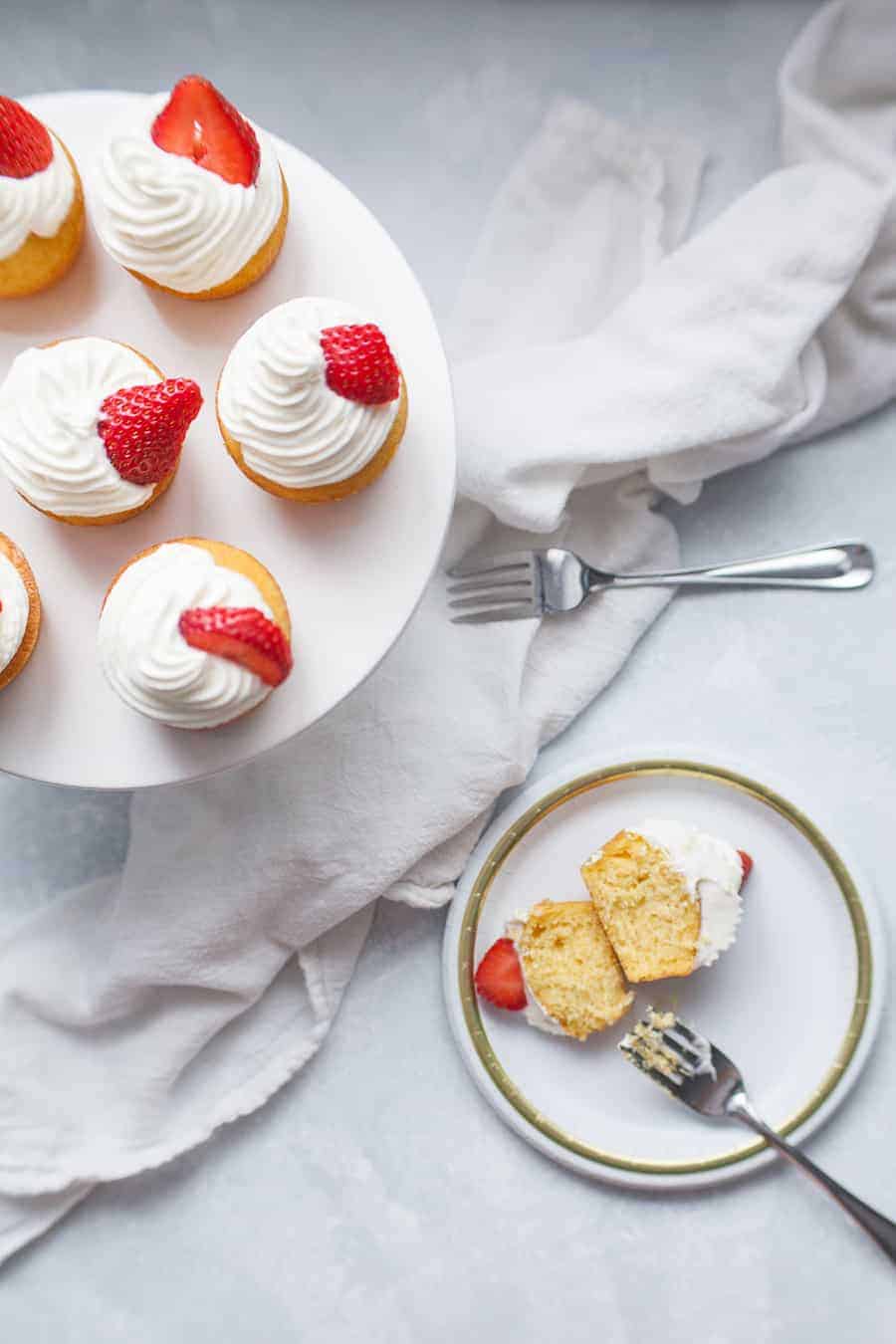 Wide photo of strawberry shortcake cupcakes with whipped cream and strawberry garnish on cupcake stand. One cupcake cut in half on small plate off to the side of the cupcake stand.