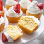 Wide photo of strawberry shortcake cupcakes with whipped cream and strawberry garnish on cupcake stand. One cupcake cut in half on cupcakes stand.