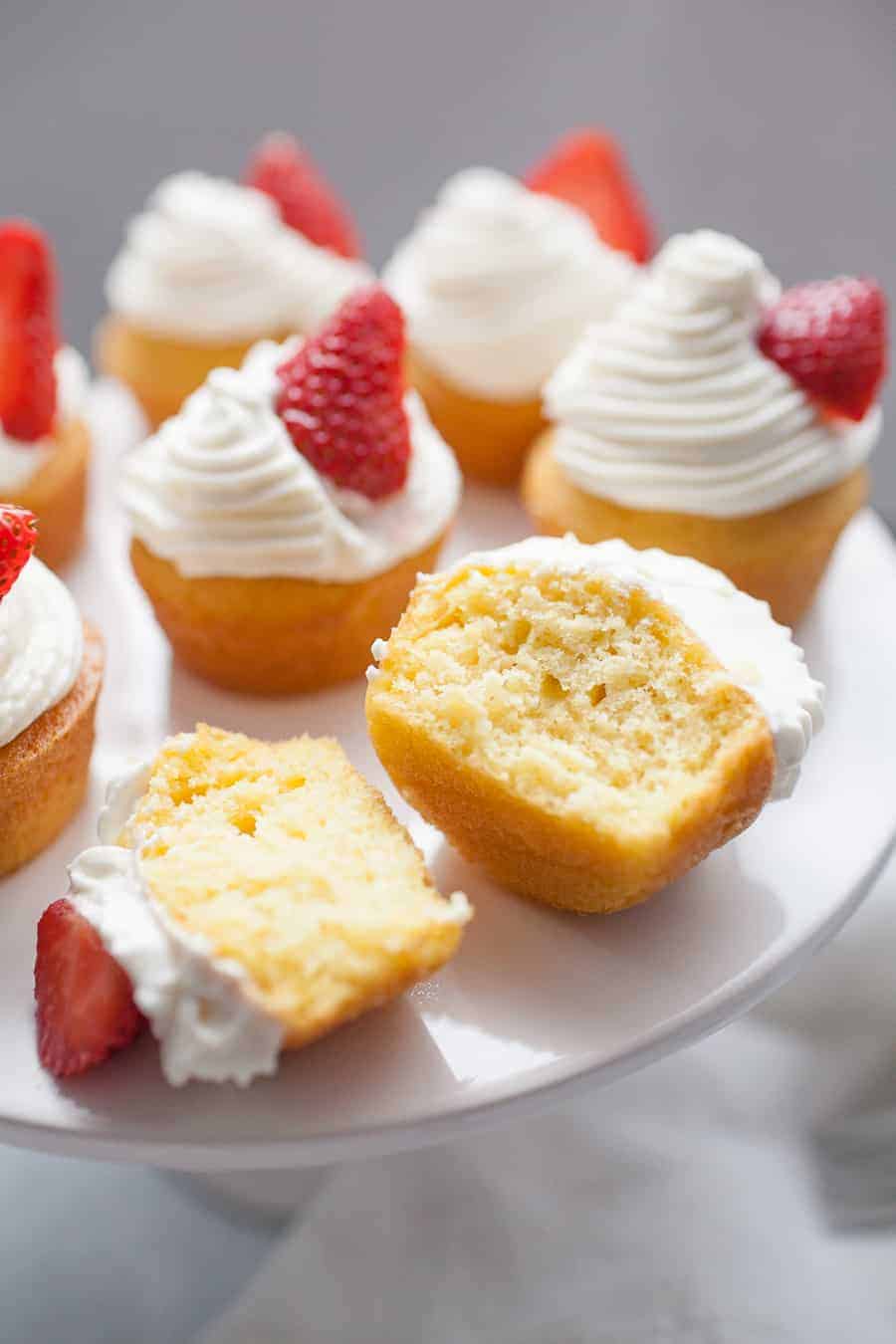 Wide photo of strawberry shortcake cupcakes with whipped cream and strawberry garnish on cupcake stand. One cupcake cut in half on cupcakes stand.