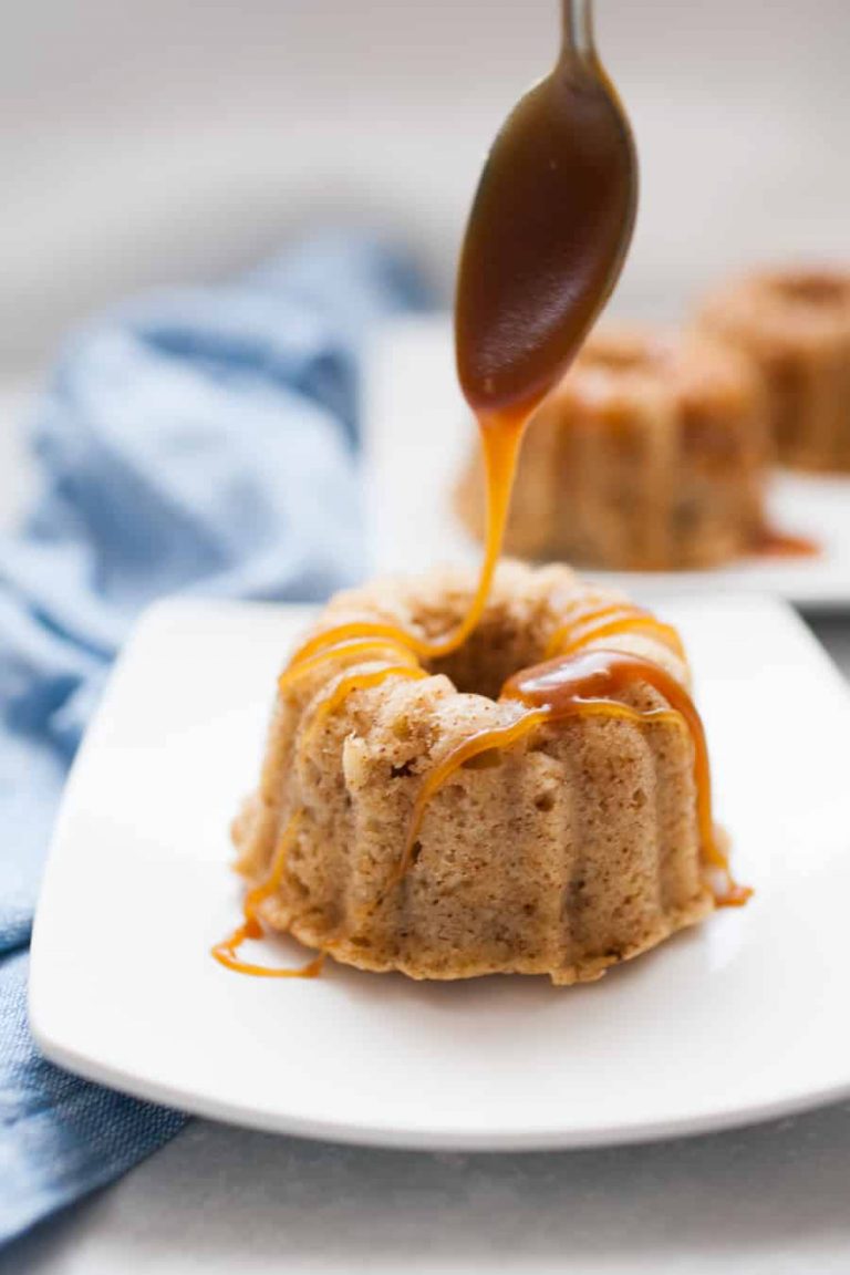 A spoon drizzles caramel sauce over a mini apple spice rum cake on a white plate, with a blue cloth in the background.