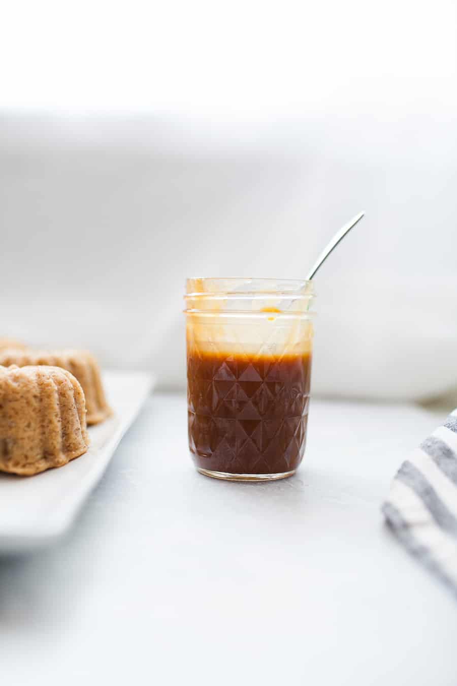 Glass mason jar of homemade boozy caramel sauce with white serving plate of apple spice cakes to the left.