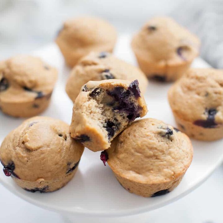 Wide photo of lemon blueberry muffins. One of them has a bite taken out of it on a cake stand.