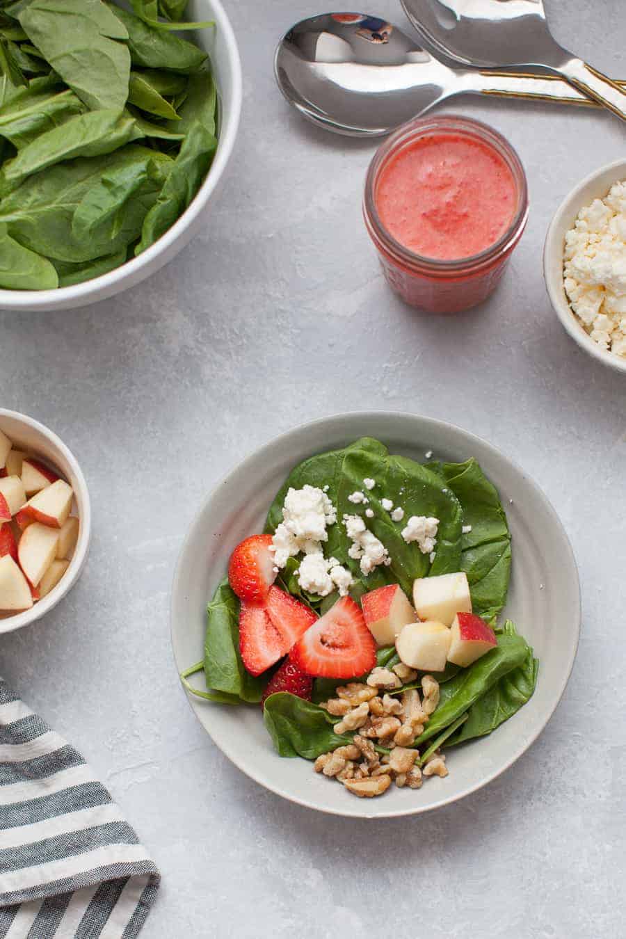 A bowl of strawberry spinach salad, surrounded by bowls of salad ingredients.