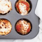 A muffin tin filled with turkey meatloaf muffins, topped with melted cheese and tomato sauce, sits invitingly on the counter. A crisp white cloth is neatly placed beside the tin.