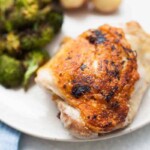 A plate with a piece of lemon pepper chicken thigh with crispy brown skin, served with broccoli and unpeeled baby potatoes in the background.