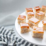Small square pieces of caramel-topped blondies and coffee creamer fudge are arranged on a white cake stand, with a striped cloth partially visible in the background.