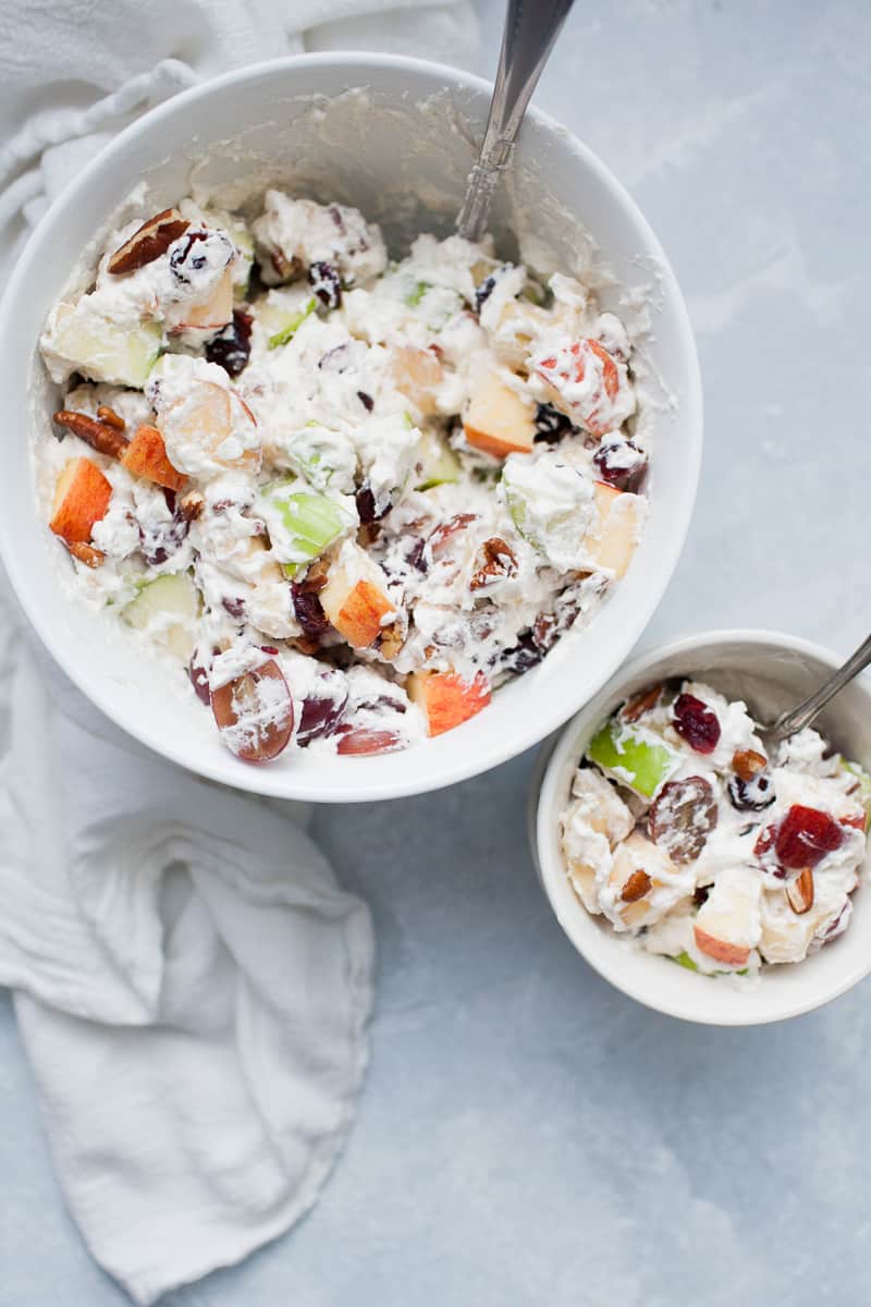 White bowls with creamy whipped cream topped fruit salad with red and green apples, grapes, bananas, cranberries and pecans.