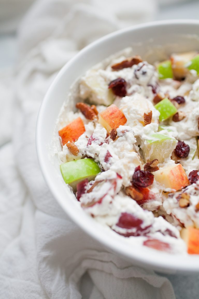 White bowl with creamy whipped cream topped fruit salad with red and green apples, grapes, bananas, cranberries and pecans.