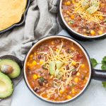 Overhead view of two bowls filled with ground beef taco soup and toppings of shredded yellow cheese and guacamole.