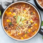 A bowl of hearty Beef Taco Soup topped with shredded cheese, avocado chunks, and black pepper. The soup contains corn, beans, tomatoes, and ground meat.