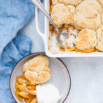 A bowl of apple cobbler with a scoop of whipped cream sits next to a baking dish of fresh peach cobbler, a spoon resting inside.