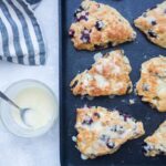 Freshly baked lemon blueberry scones with icing are arranged on a black baking tray, with a jar of glaze and a spoon nearby on a white surface.