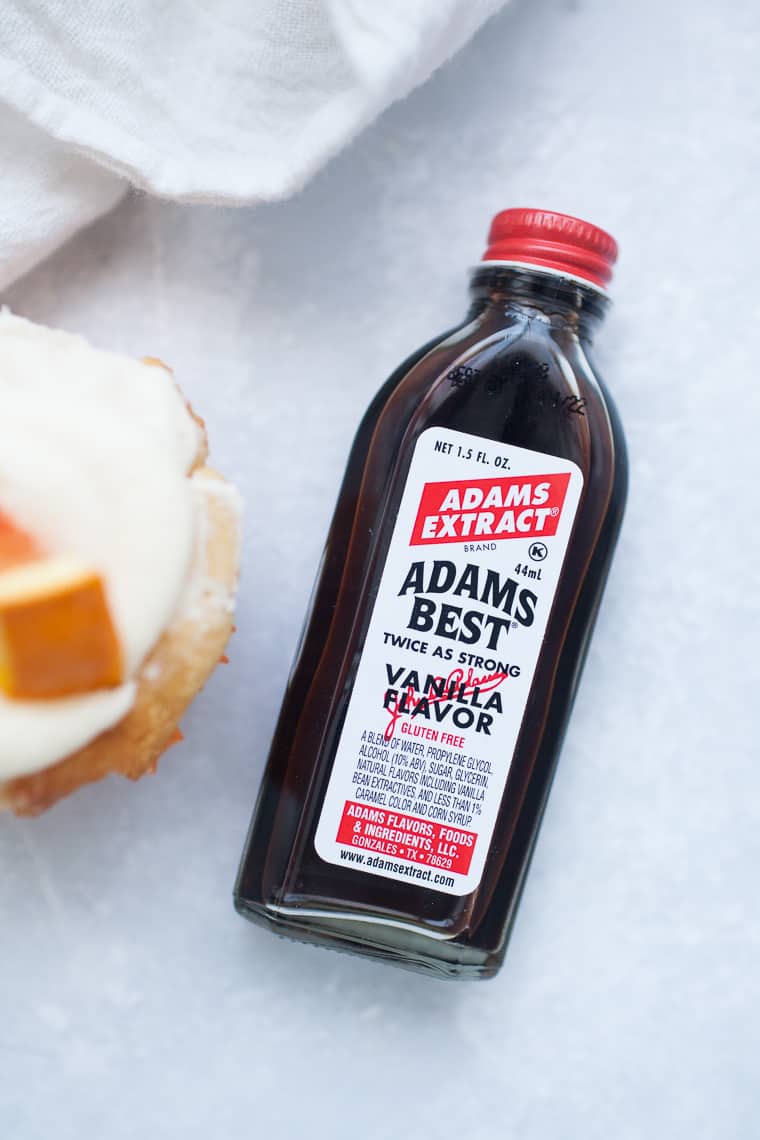 A 1.5-fl-oz bottle of Adams Best Vanilla Flavoring rests on a white surface next to a partially visible dessert topped with cream and fruit, perfect for flavoring your orange cupcakes.