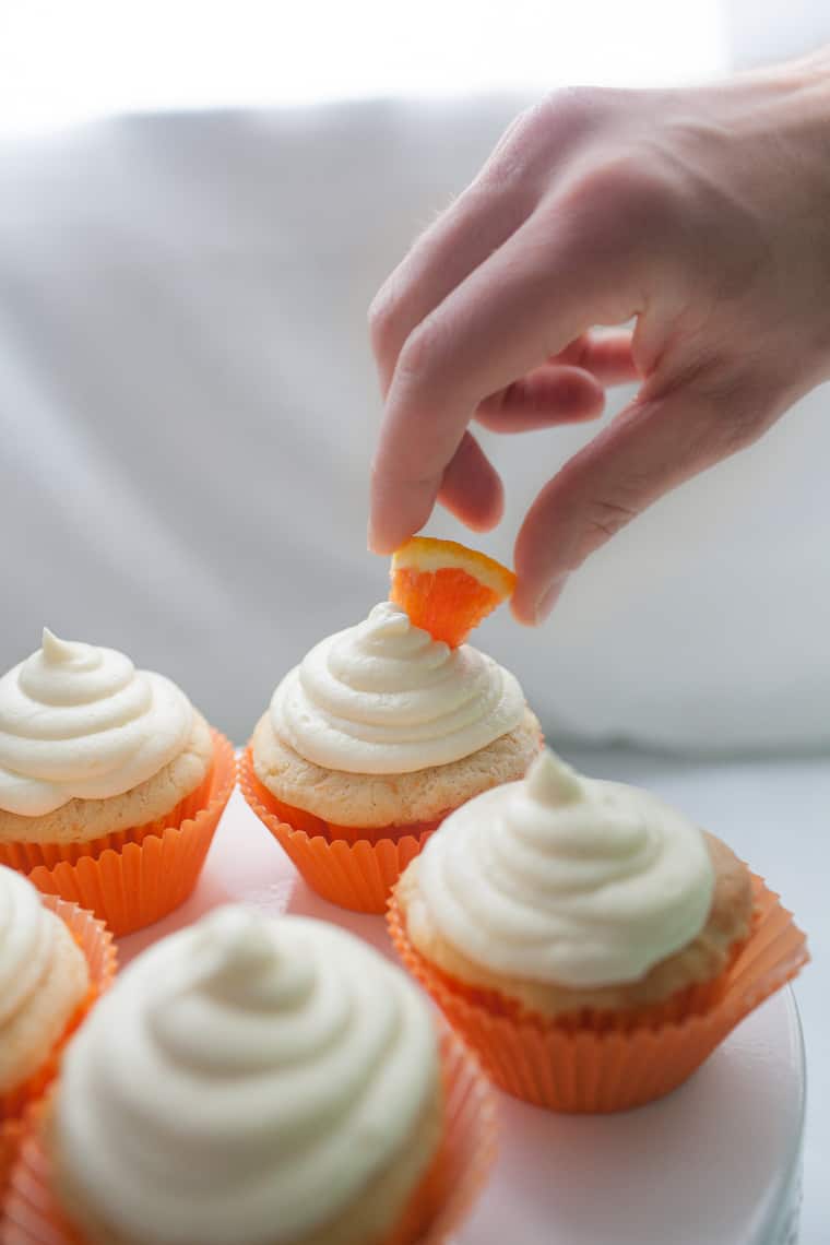 A hand placing an orange slice atop a cupcake with white frosting, surrounded by other delectable orange cupcakes in matching wrappers.