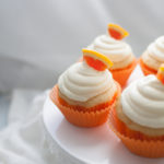 Four orange cupcakes with white frosting and orange slice garnishes, placed in vibrant orange wrappers, arranged on a pristine white cake stand.