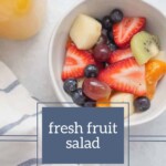 A bowl of Fresh Fruit Salad brimming with strawberries, blueberries, grapes, kiwi, and melon rests on a light surface beside a striped towel.
