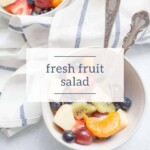 A bowl of Fresh Fruit Salad with strawberries, grapes, kiwi, blueberries, orange slices, and apple sits next to a striped towel and two spoons.