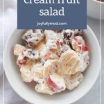 A bowl of fruit salad with whipped cream, featuring sliced bananas, strawberries, and grapes. Text overlay reads "whipped cream fruit salad joyfullymad.com." Indulge in the sweetness of fresh fruits that pairs perfectly with our ginger molasses snickerdoodles.