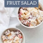 Two bowls of whipped cream fruit salad on a light surface, with mixed fruit pieces. The text "whipped cream FRUIT SALAD" and "joyfullymad.com" is displayed on the image. A plate of ginger molasses snickerdoodles adds a touch of warmth to this delightful scene.
